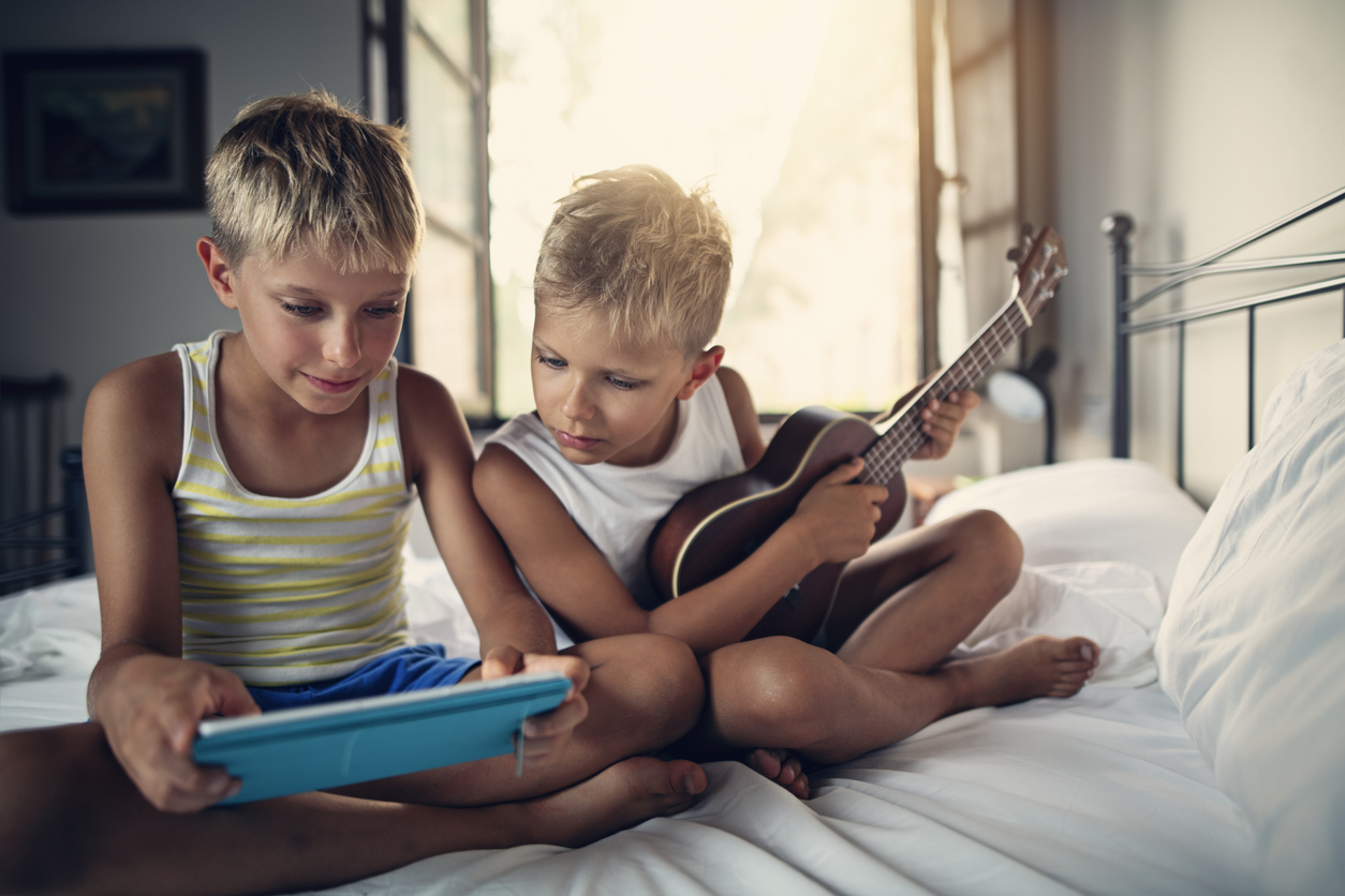 A preschool boy playing a ukulele while his brother uses a tablet